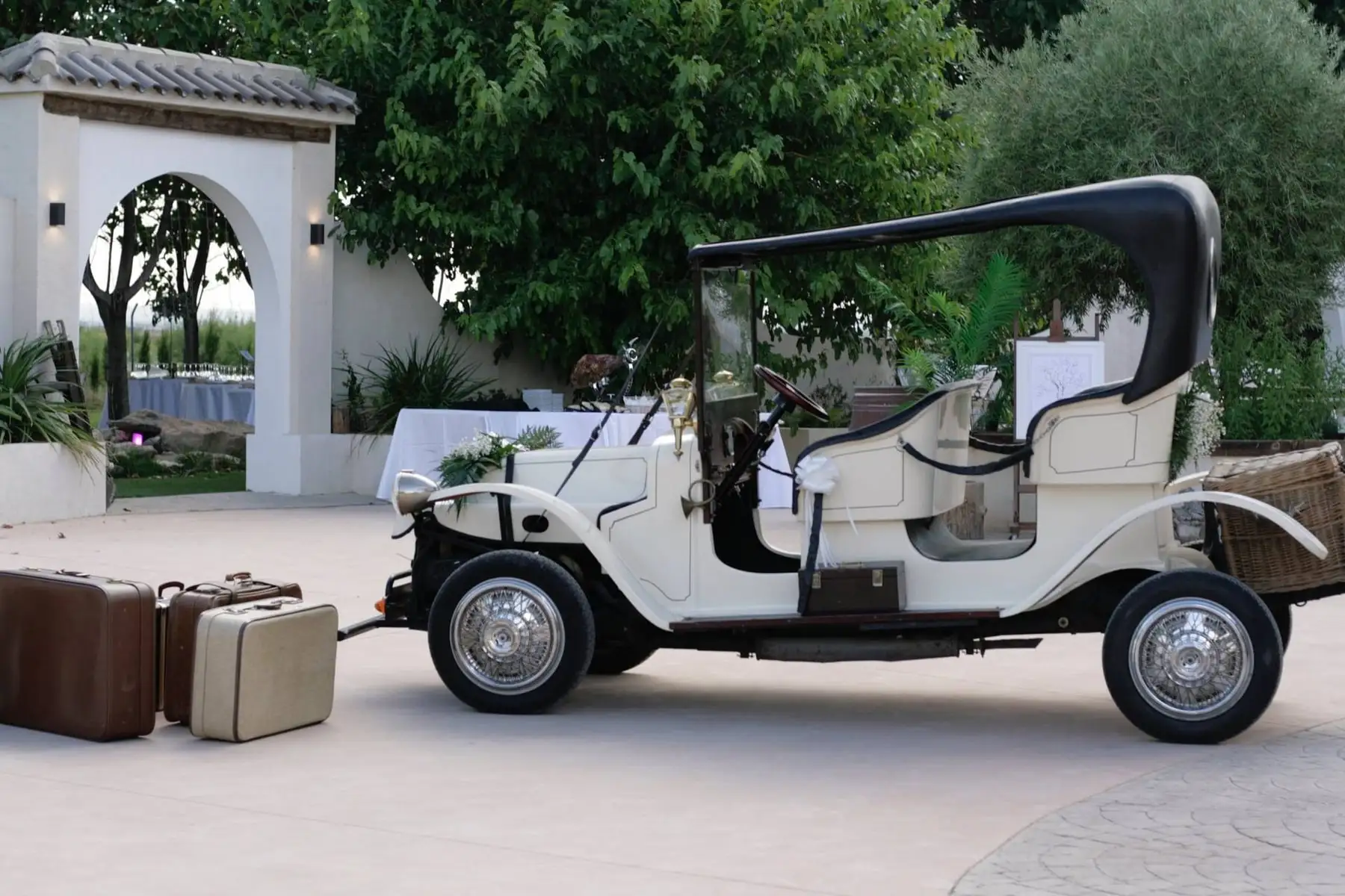 Un coche antiguo de color crema con maletas de cuero, delante de un arco blanco con tejadillo de teja