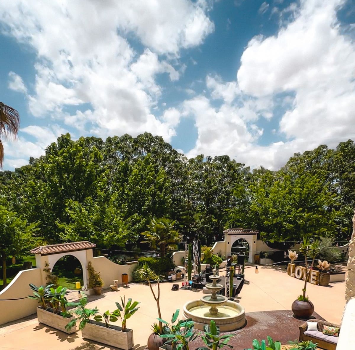 El patio de entrada de Finca Los Prados visto desde arriba, con su fuente en el centro, arcos blancos con tejadillo de teja y una arboleda detrás bajo un cielo de nubes